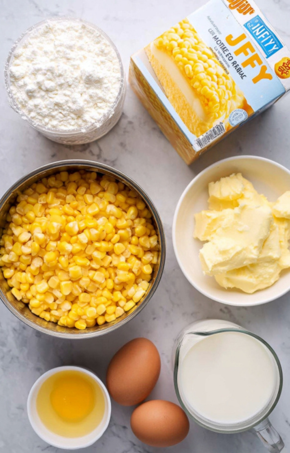 All the ingredients for Corn Bake Casserole Jiffy: canned corn, Jiffy mix, butter, eggs, milk, and flour arranged on a marble counter, ready for quick assembly.