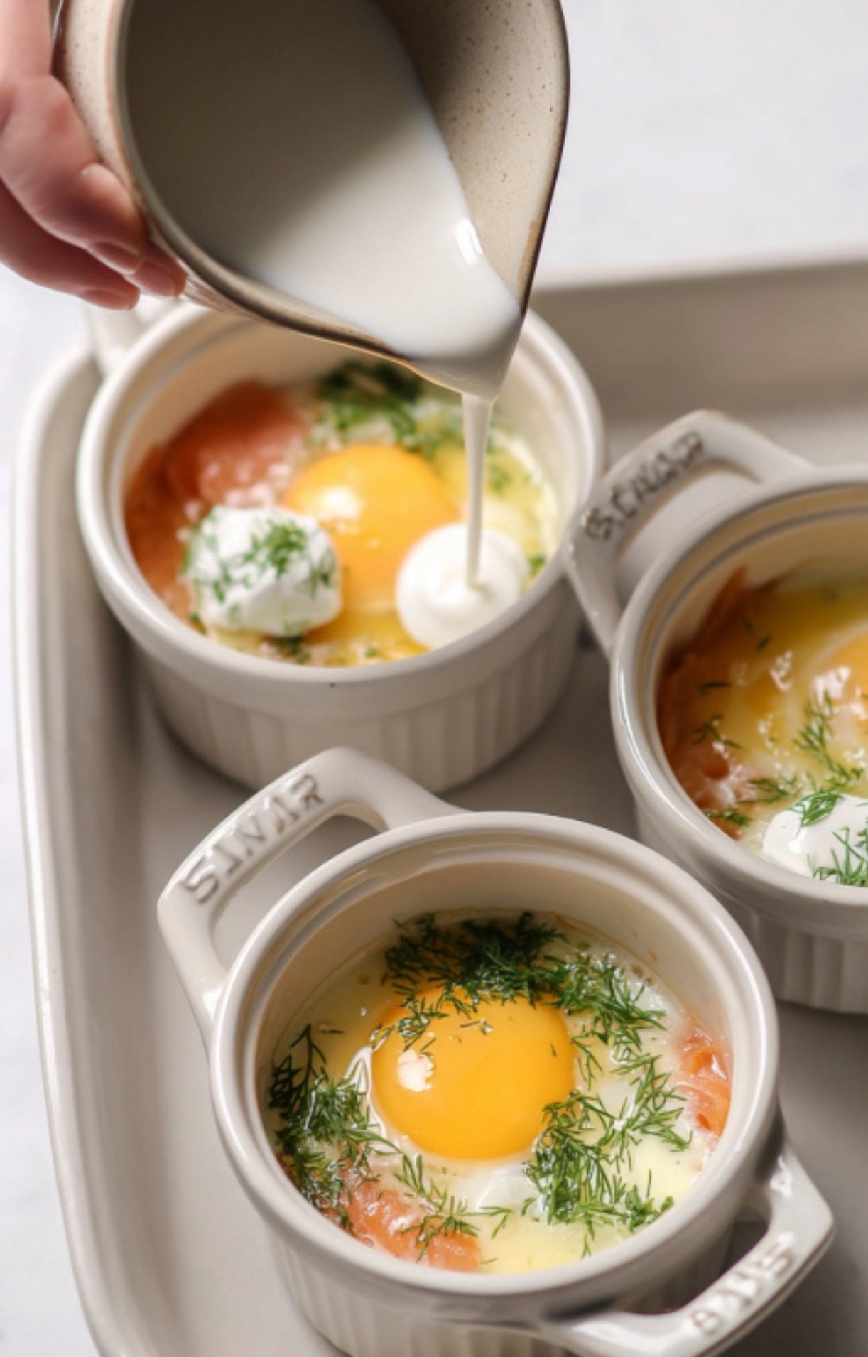 Cream being poured into cocottes with eggs, salmon, and dill, before baking in the oven for a creamy brunch.