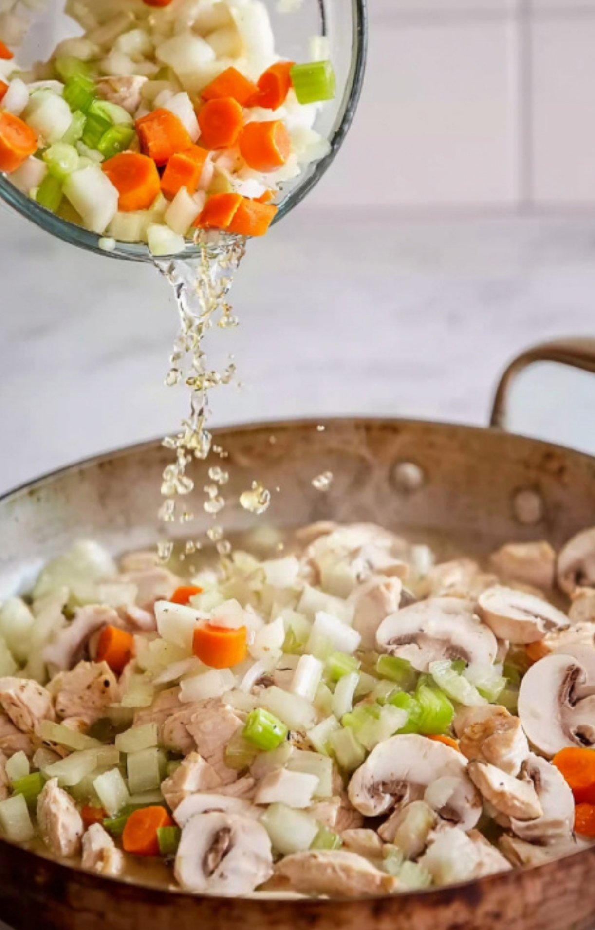 Pouring chopped carrots, celery, onions, and broth into a skillet with mushrooms and chicken for fricassee.