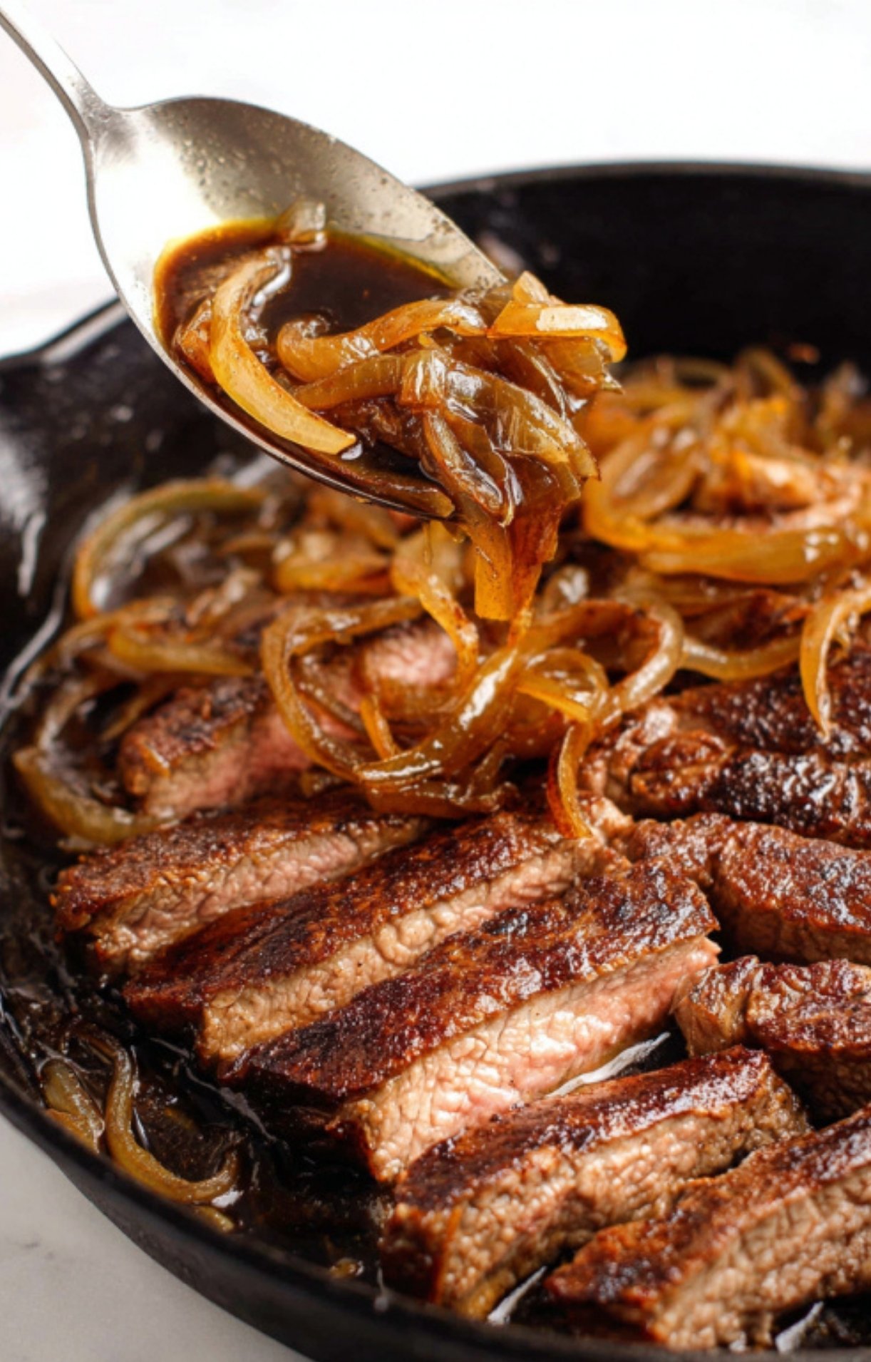 Sliced steak strips in a cast iron skillet topped with caramelized onions and savory pan sauce.