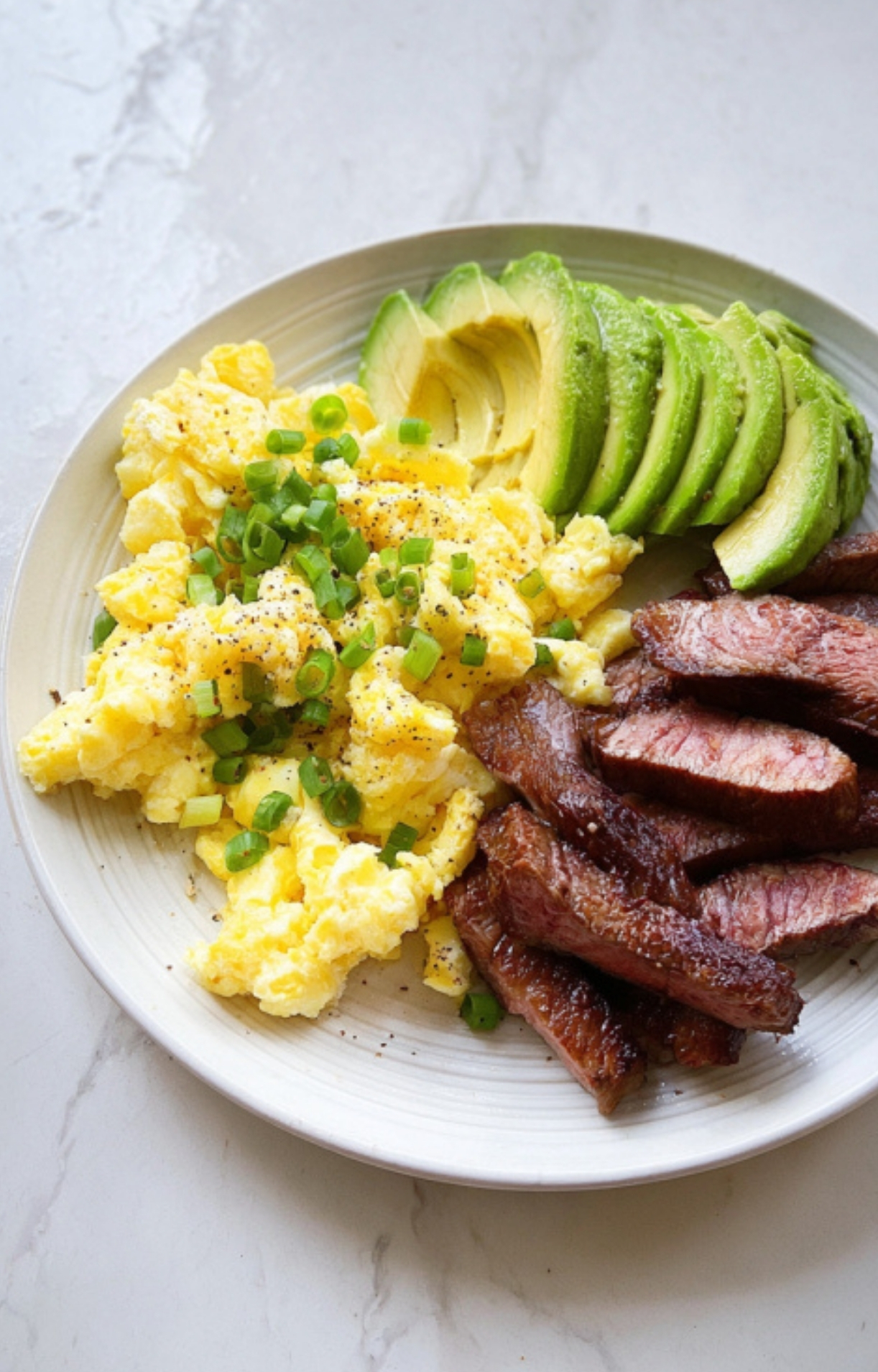 Steak strips, fluffy scrambled eggs, and sliced avocado on a plate with green onions and black pepper.