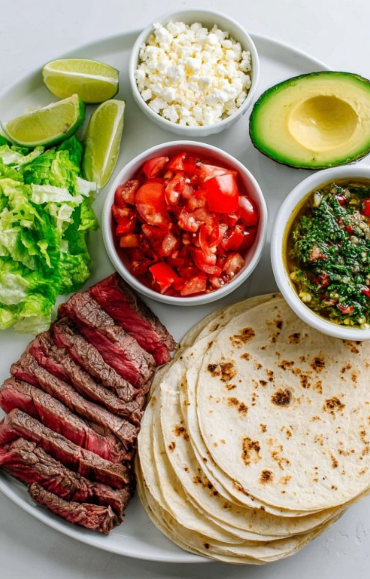 DIY shaved steak taco bar with tortillas, sliced steak, romaine lettuce, tomatoes, feta cheese, avocado, and chimichurri sauce, ideal for parties, Taco Tuesday, or Cinco de Mayo.