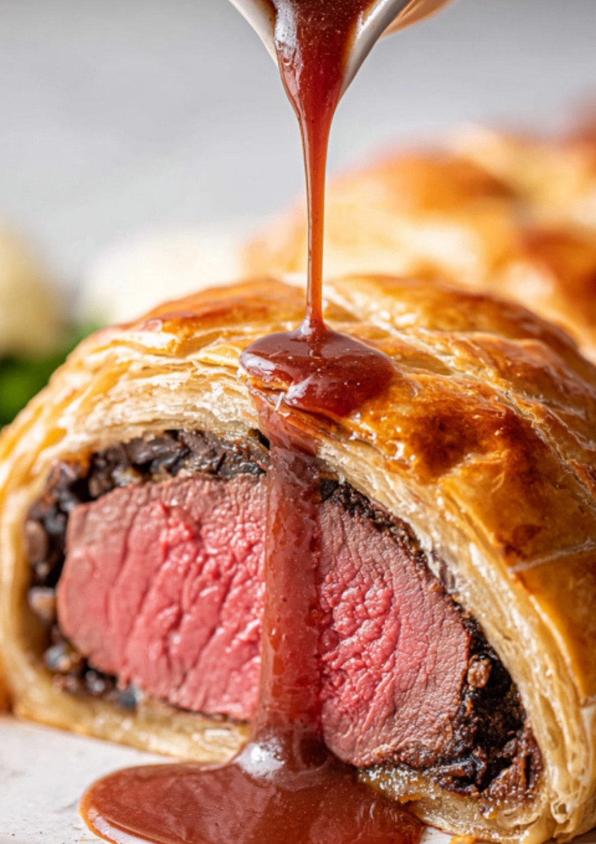 Red wine sauce being poured over a sliced beef wellington with a tender pink center and crisp pastry, a festive main course.