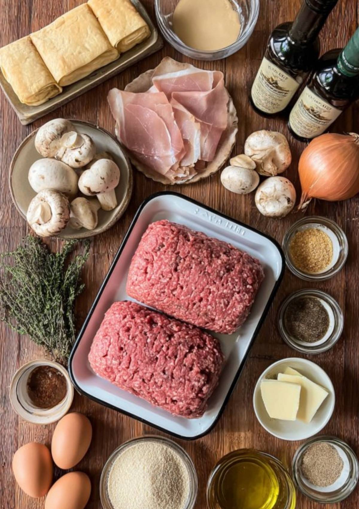 Flatlay showing ground beef, puff pastry, prosciutto, fresh mushrooms, eggs, seasonings, and herbs. All the ingredients you need to make homemade mini beef wellingtons for cozy dinners and special occasions.