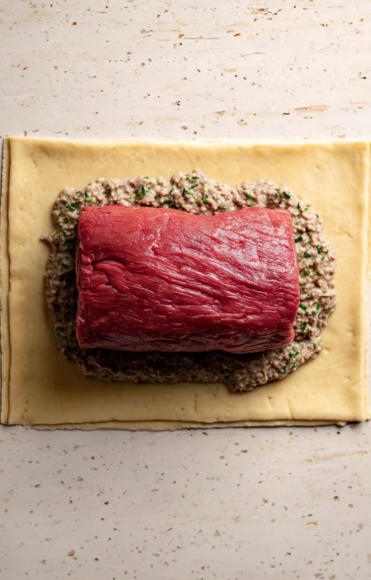 Beef tenderloin placed on a layer of mushroom duxelles spread over puff pastry, showing an important step in assembling classic beef wellington. Great for holiday dinners and entertaining.