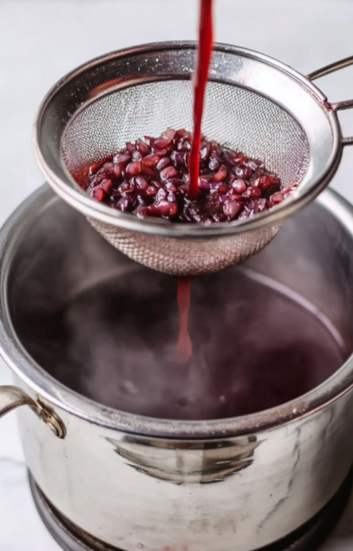 Pouring red wine reduction through a fine mesh strainer, showing a key step for making smooth beef wellington sauce at home.