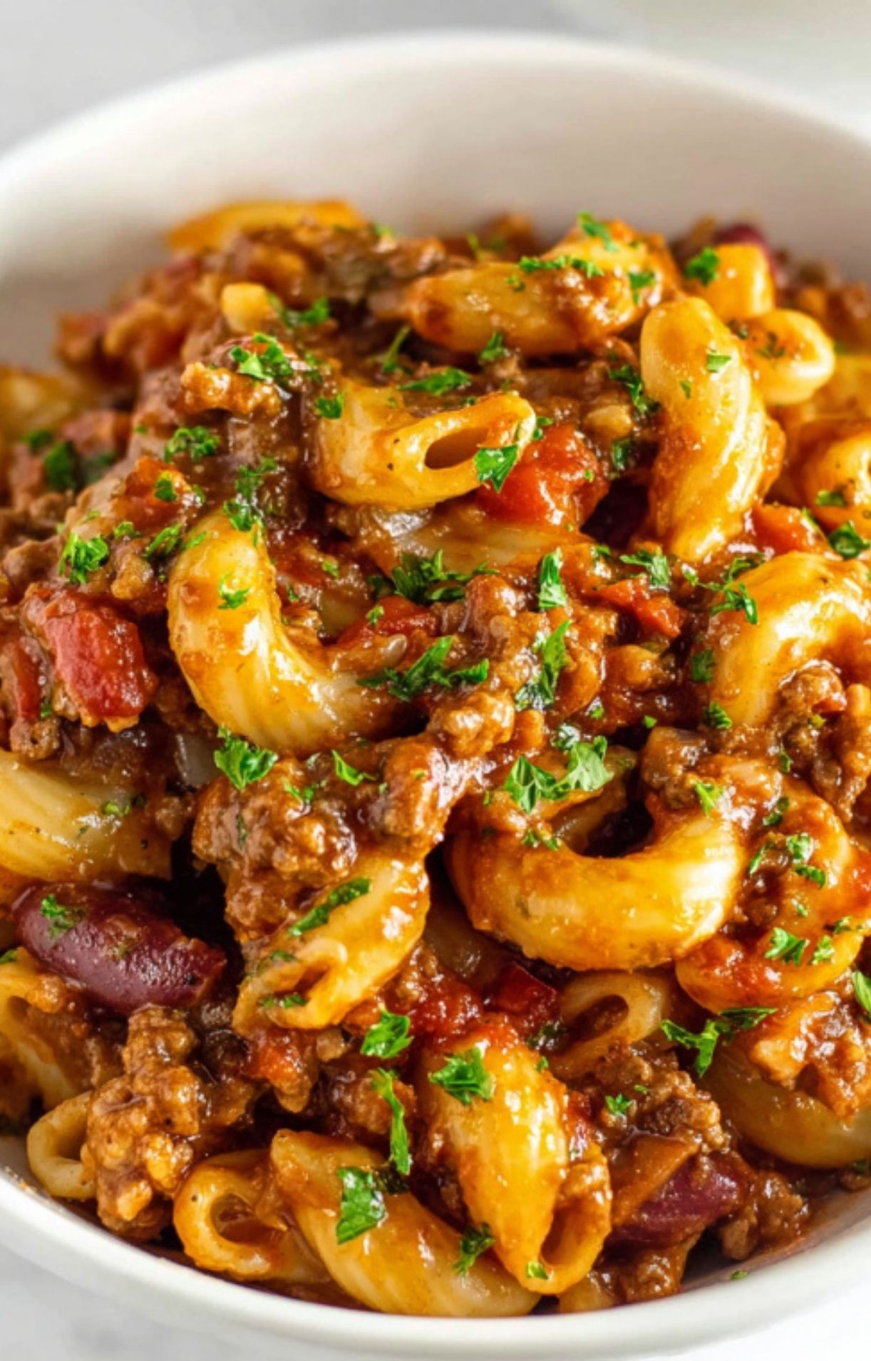 Bowl of hot, cheesy goulash with beef, pasta, and beans, garnished with parsley. Quick dinner idea that’s kid-approved and filling.