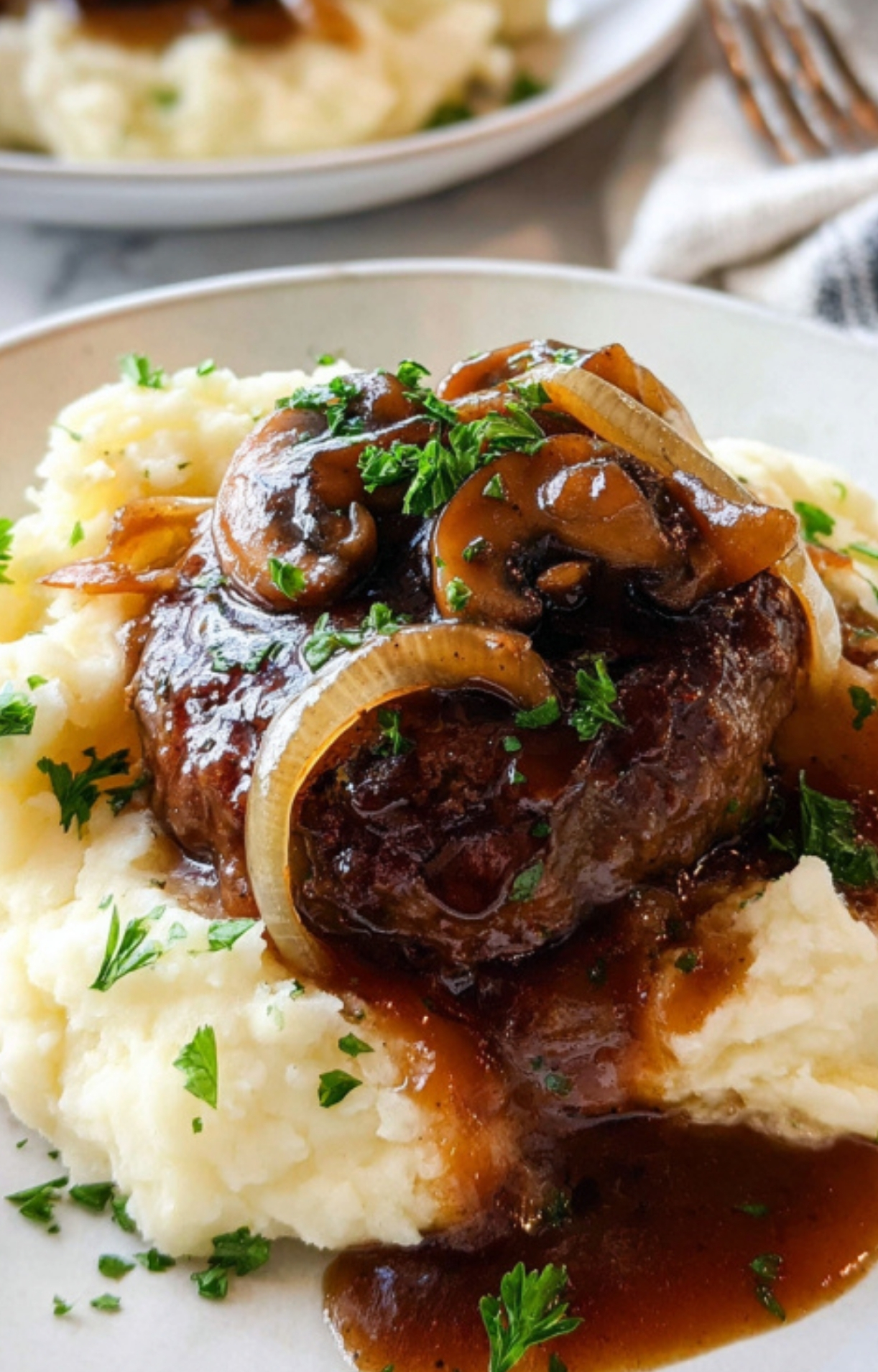 Plate of Salisbury steak topped with mushroom gravy, served over creamy mashed potatoes and sprinkled with fresh herbs.