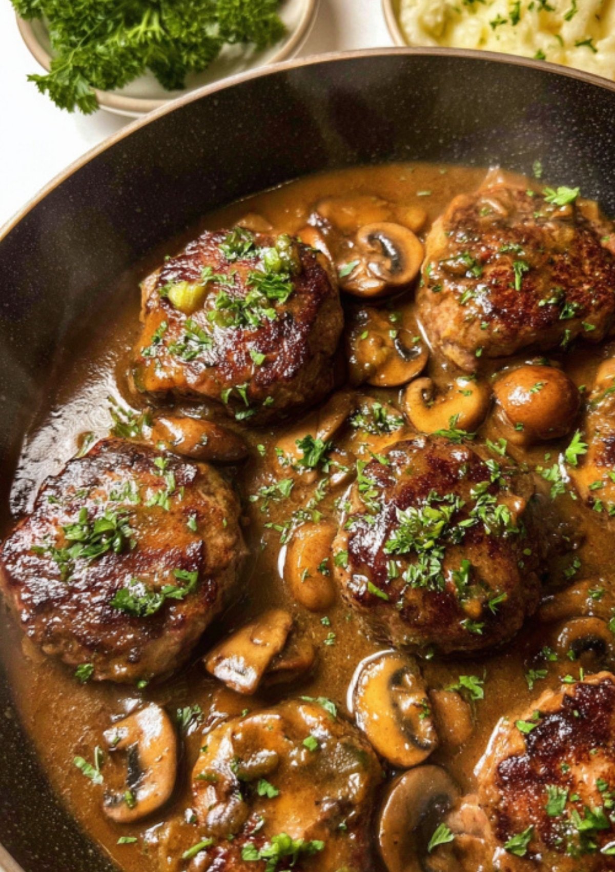 Hearty beef patties in a skillet, smothered in mushroom gravy and fresh herbs.