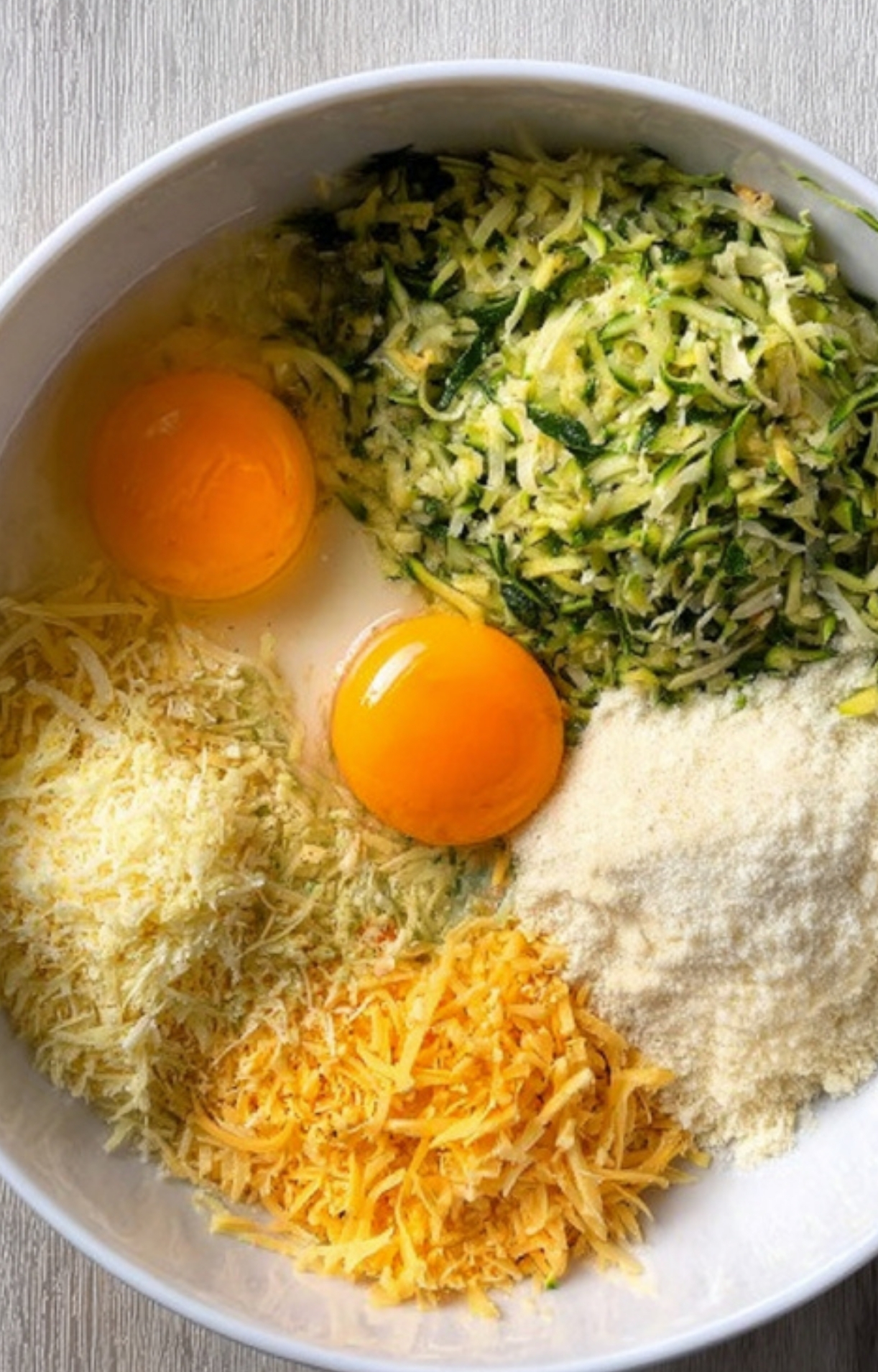Bowl of shredded zucchini, cracked eggs, parmesan, cheddar, and mozzarella ready to mix for casserole