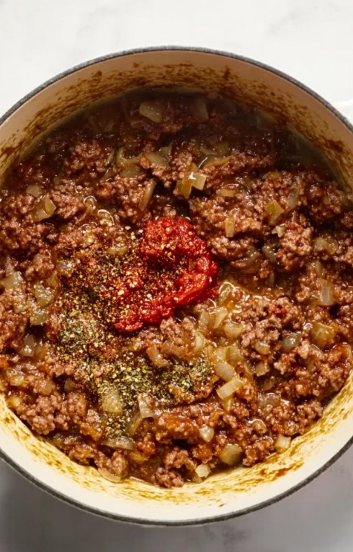 Ground beef, onions, and tomato paste with herbs and spices cooking in a large pot. Step for building rich flavor in cheesy American goulash.