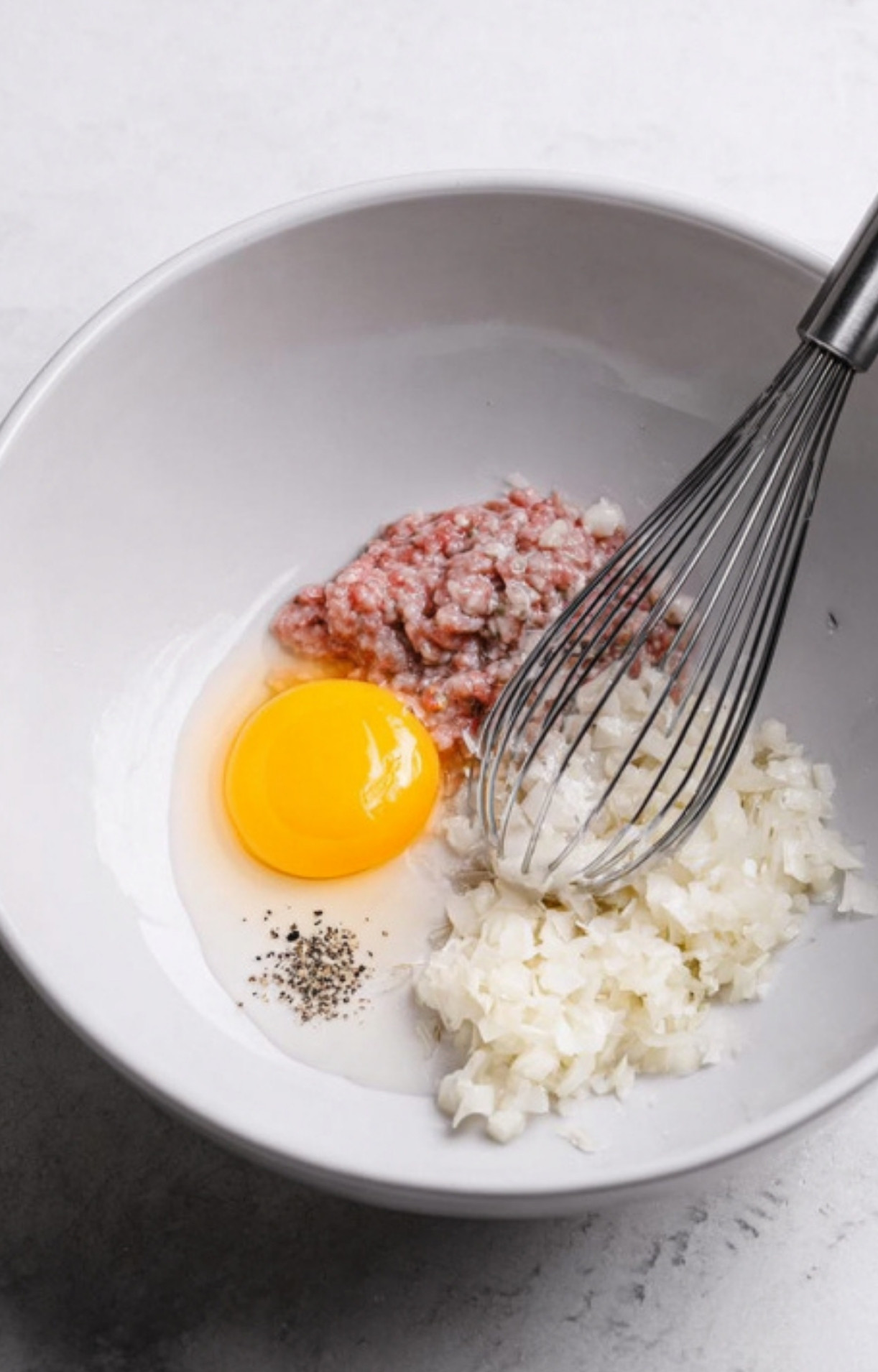Bowl with ground beef, fresh chopped onion, egg, and pepper, ready to be mixed for a classic ground beef Wellington.