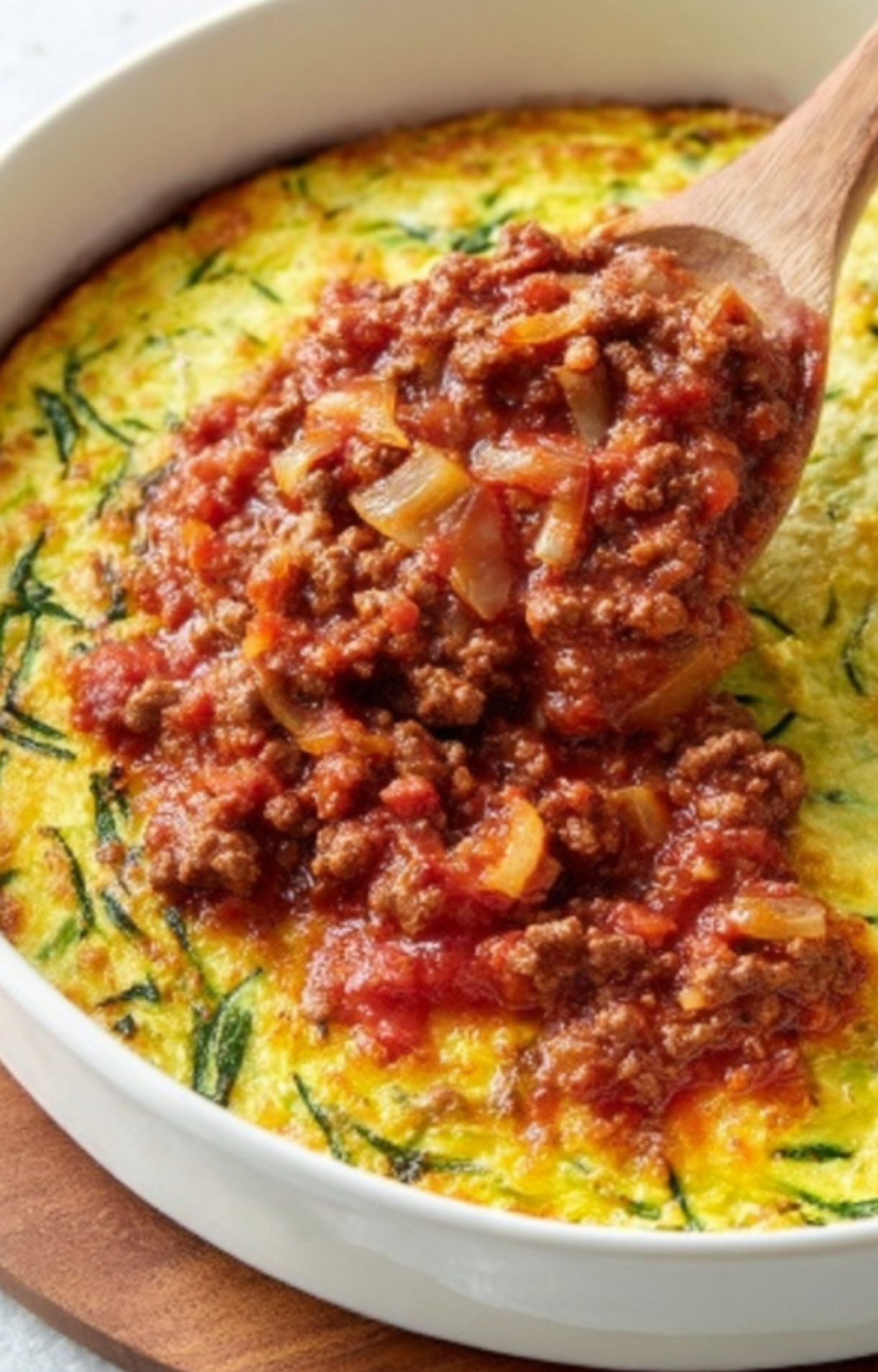 Tomato beef sauce with onions being spread over a baked zucchini and egg casserole base