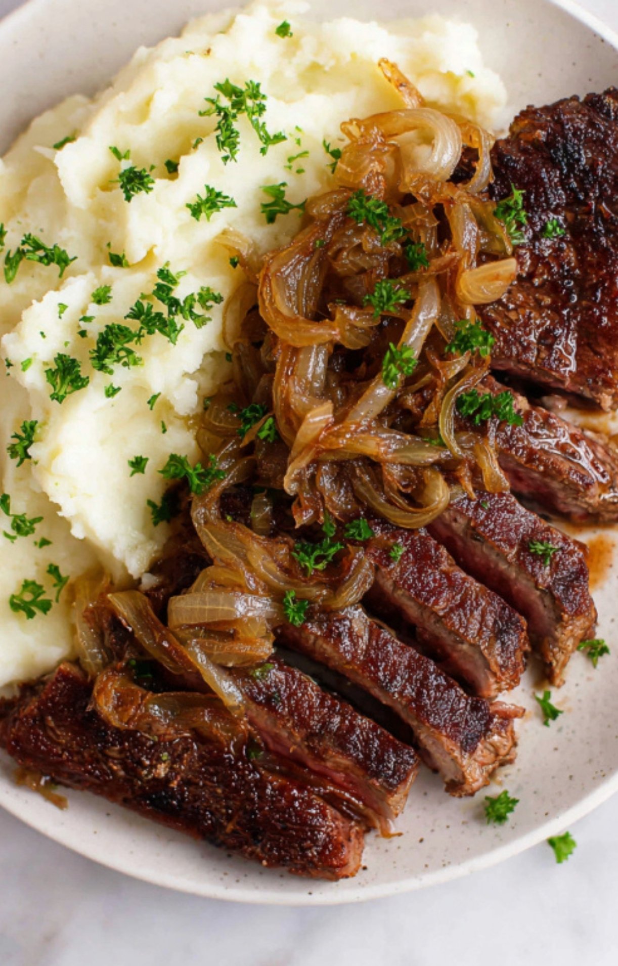 Sliced steak strips topped with buttery caramelized onions, served alongside creamy mashed potatoes and fresh parsley.