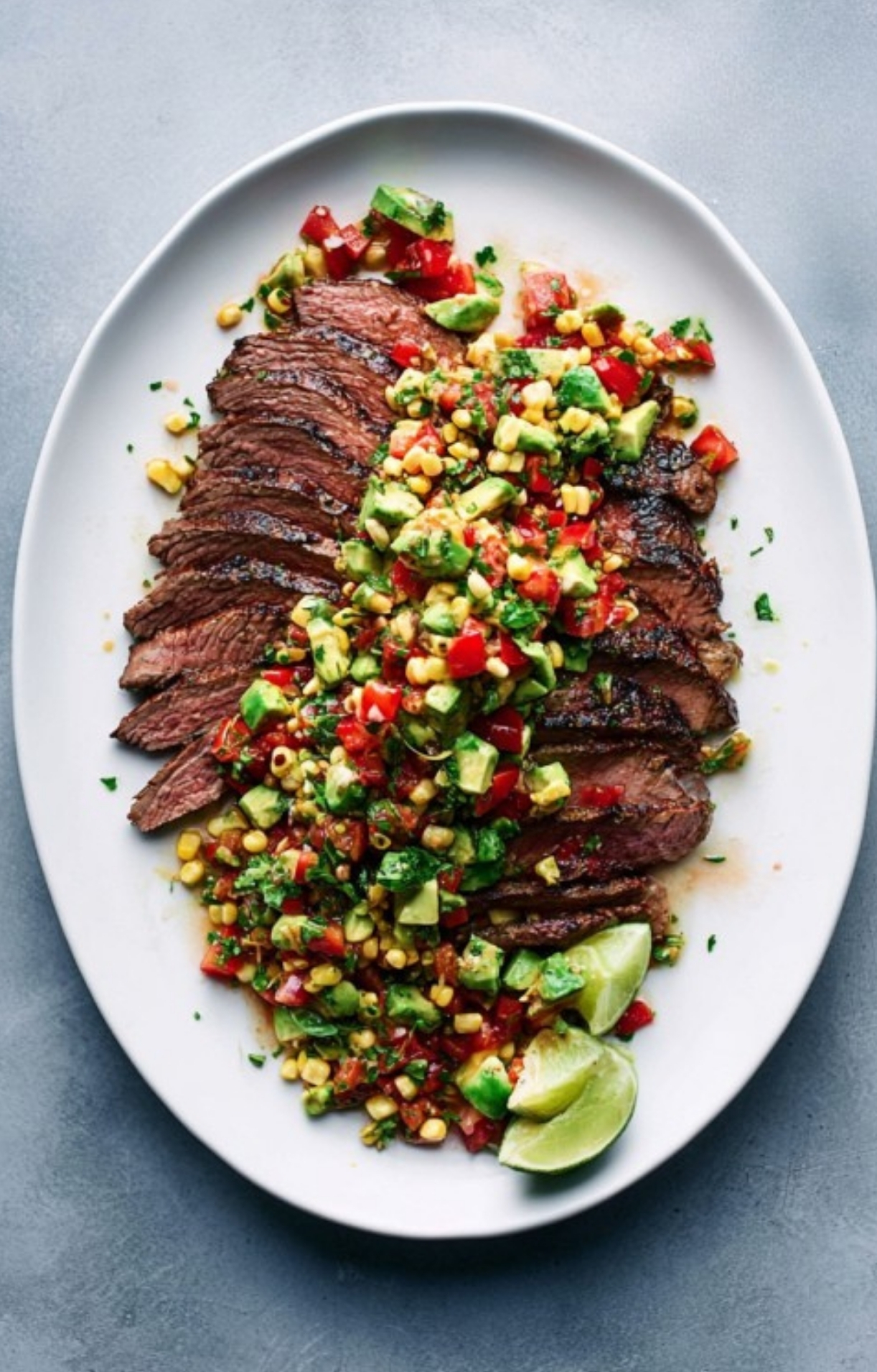Sliced grilled flank steak topped with fresh avocado corn salsa and lime wedges, served on a white platter.