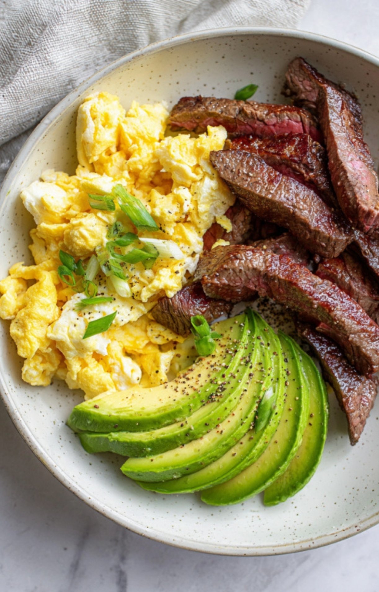 A bowl filled with leftover steak strips, scrambled eggs, and avocado slices for a filling morning meal.
