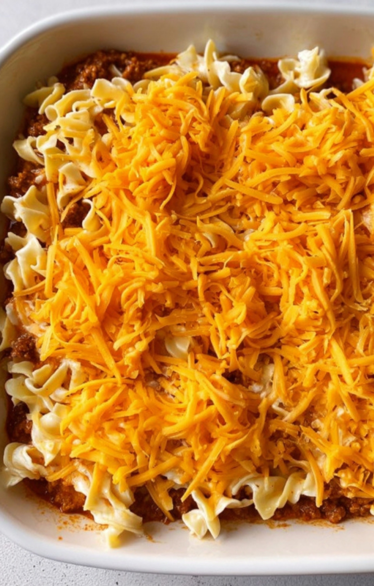 Closeup of casserole layered with tender noodles, creamy sauce, and topped with bright cheddar cheese, ready to bake for a comforting weeknight meal.