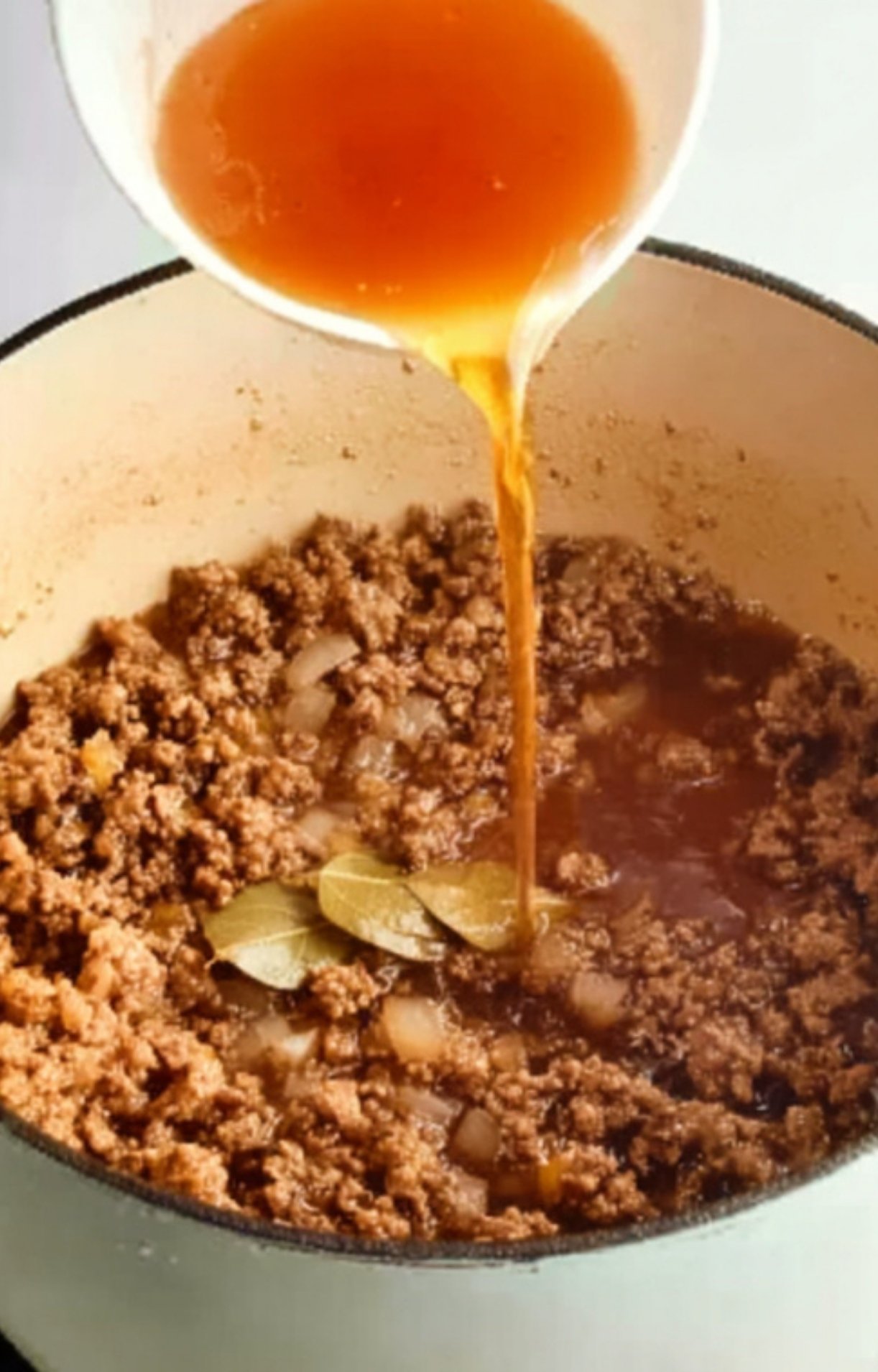 Pouring beef broth into browned beef and onions in a Dutch oven, building layers of flavor for classic goulash.