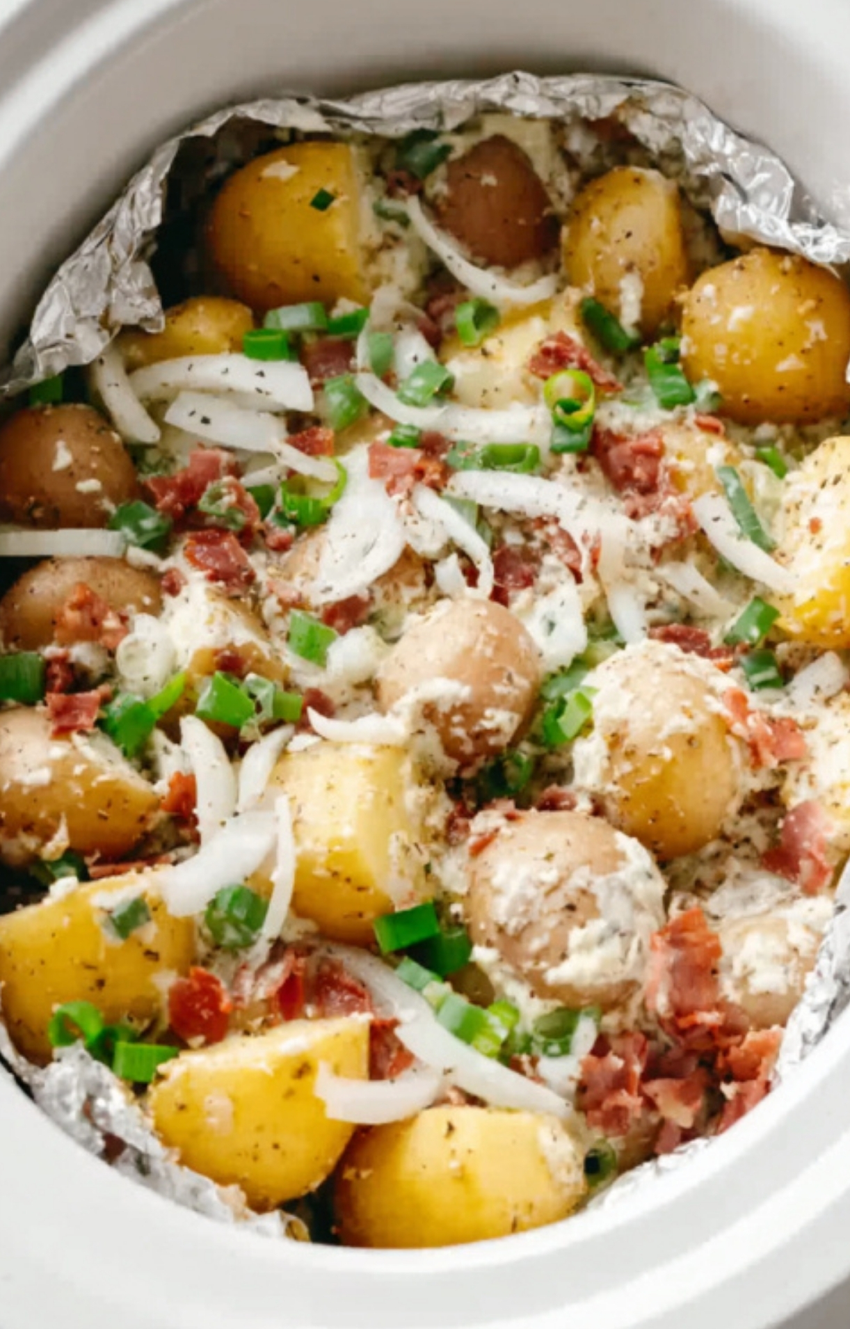 Potatoes, onions, and bacon layered in foil inside a crock pot, ready to become creamy cheesy ranch potatoes for an easy slow cooker dinner.