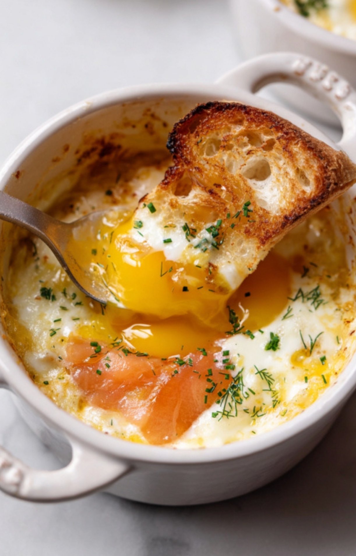 Ramekin filled with baked egg, smoked salmon, and chopped herbs, with toast dipped into a soft, golden yolk.