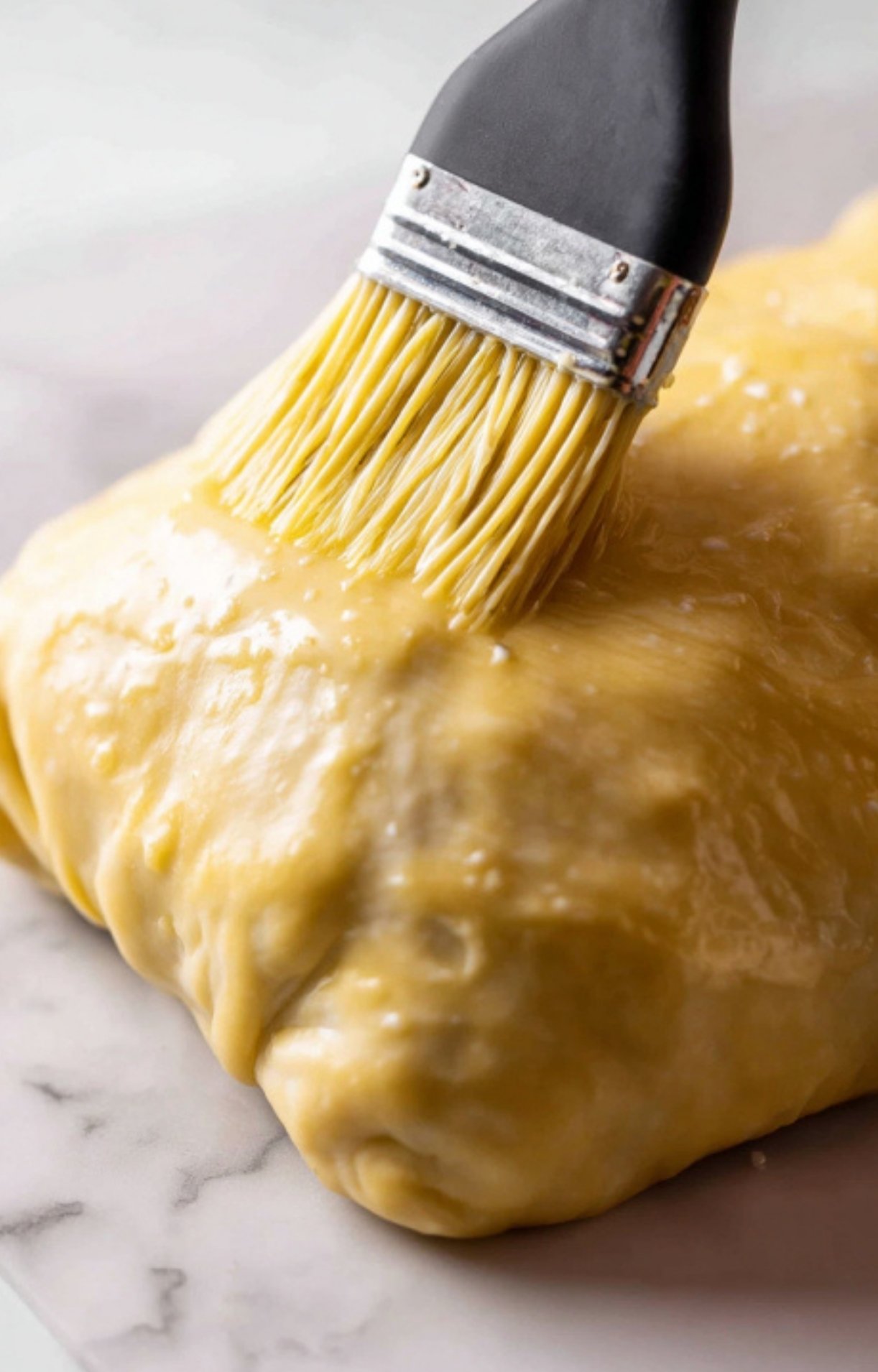 Close-up of a pastry brush applying egg wash to puff pastry-wrapped beef tenderloin, an essential step for a crisp and shiny wellington.