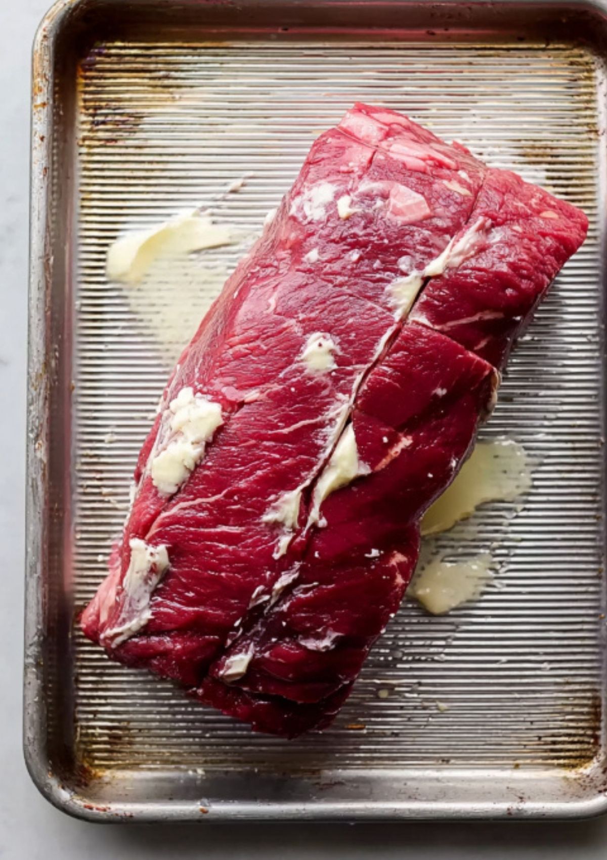 Raw beef tenderloin rubbed with butter, ready to roast for a simple beef wellington recipe, shown on a metal baking tray.