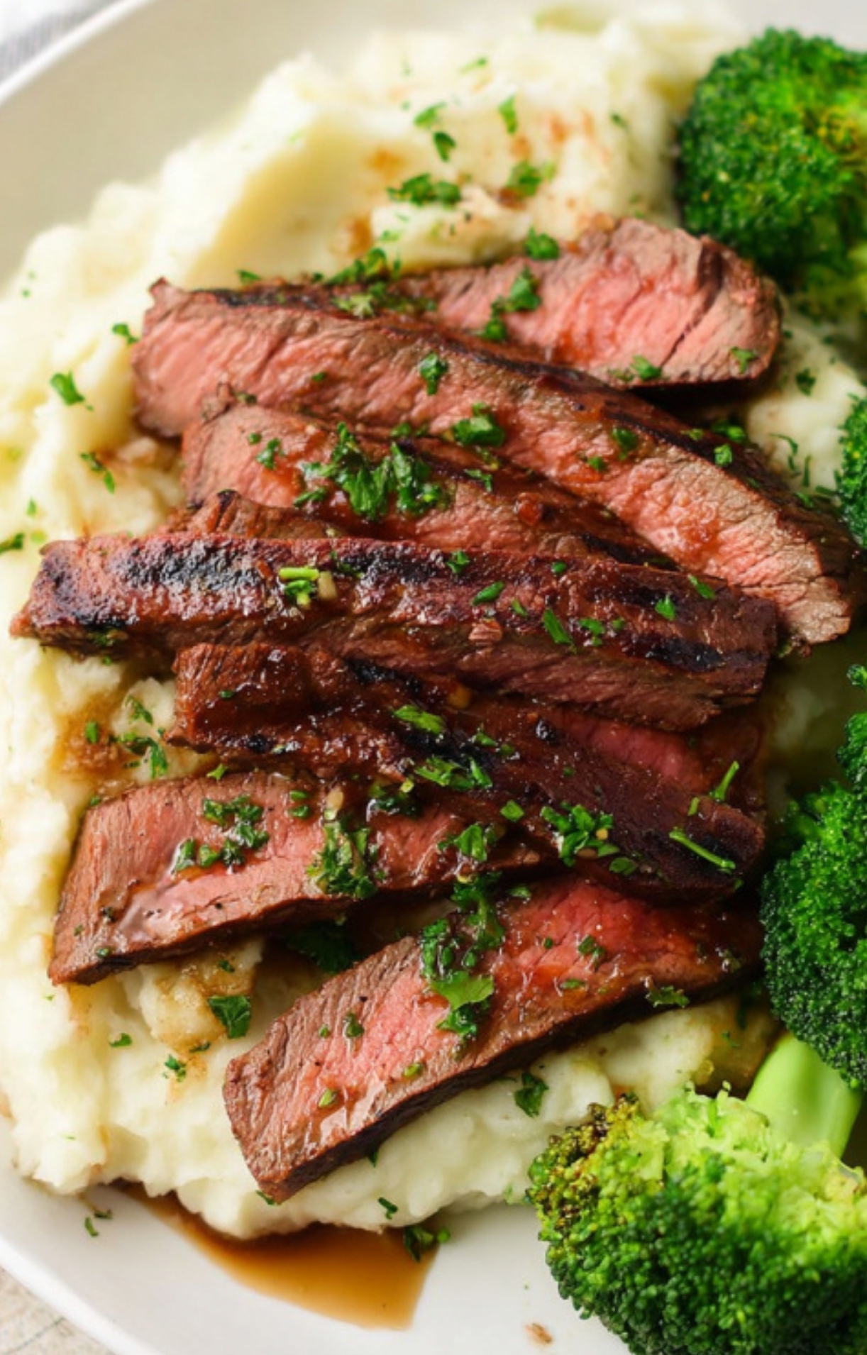 Sliced marinated flank steak served over creamy mashed potatoes with broccoli, garnished with fresh herbs for a balanced, high-protein meal.