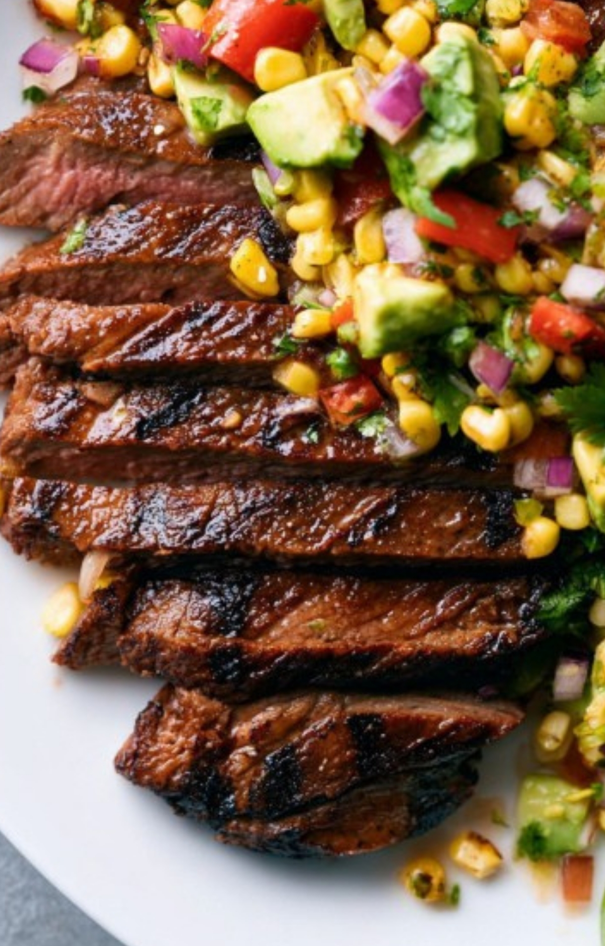 Sliced grilled flank steak topped with fresh avocado corn salsa on a white plate, perfect for a healthy summer dinner.