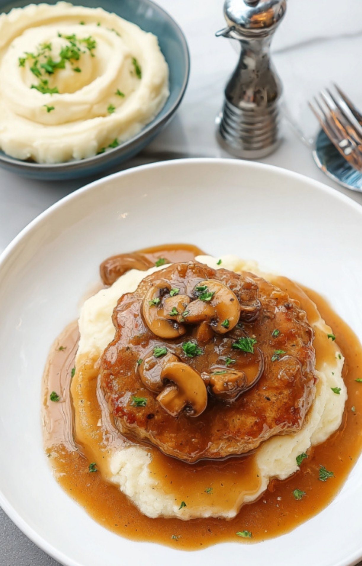 Plate of ground turkey Salisbury steak topped with mushroom gravy served over creamy mashed potatoes, with a bowl of mashed potatoes and pepper mill in the background.