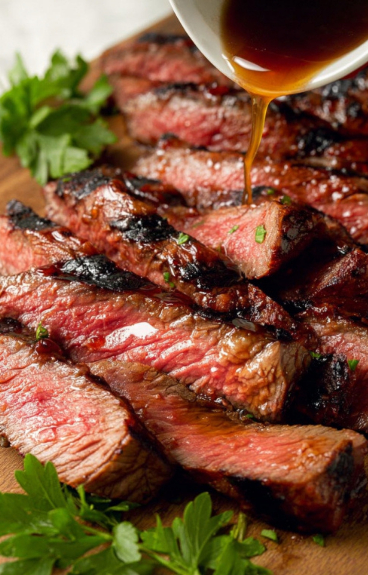 Sliced marinated flank steak on a cutting board, topped with a drizzle of steak sauce, garnished with parsley for a crowd-pleasing barbecue dish.
