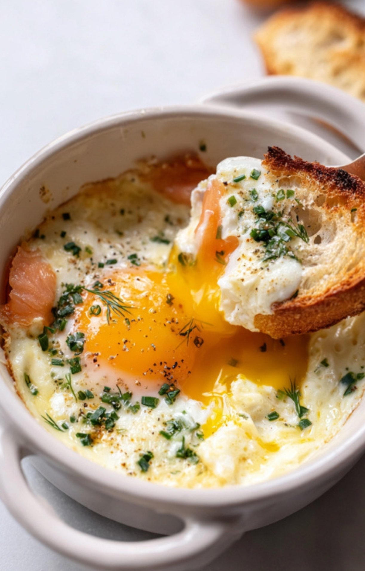 A soft-baked egg in a ramekin with smoked salmon and herbs, yolk running as toasted bread is dipped inside.