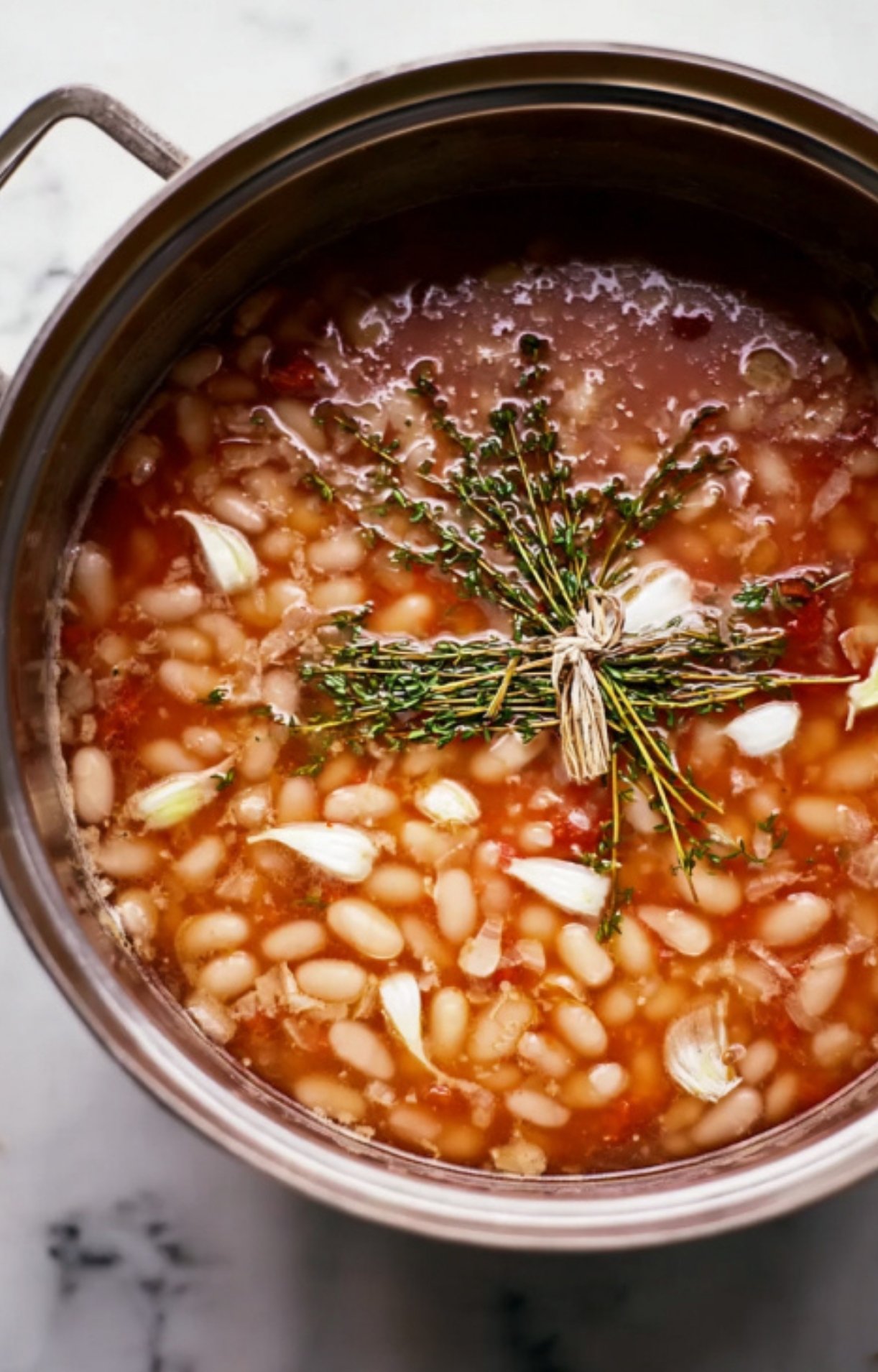 Pot of beans simmering with garlic and a bundle of fresh thyme, the aromatic base for a cassolette recipe, essential for capturing authentic French flavor in every bite.