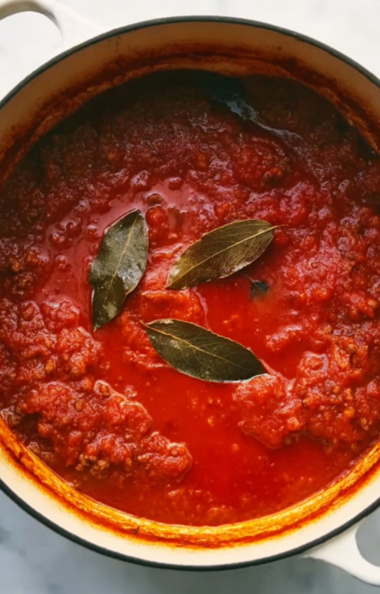 Dutch oven with homemade tomato sauce and bay leaves simmering. Flavorful base for cheesy beef goulash, great for cozy fall and winter meals.