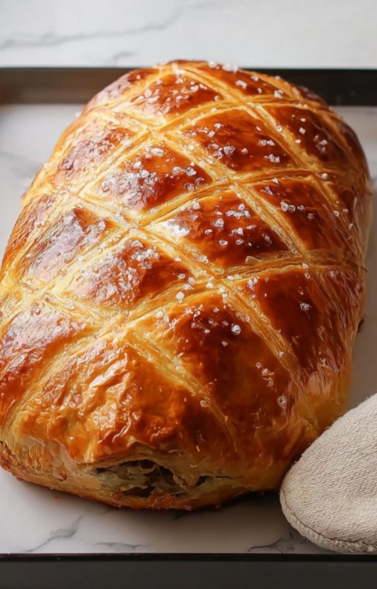 Whole sous vide beef wellington with a golden flaky crust and sea salt on top, cooling on a tray and ready for slicing.