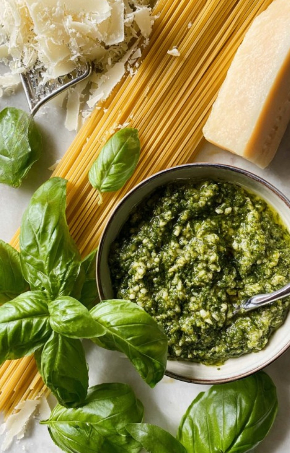 Flat lay of fresh basil leaves, a bowl of homemade pesto, uncooked spaghetti, and a wedge of parmesan cheese, showcasing the simple ingredients for an easy week night dinner recipe with Italian flavors.