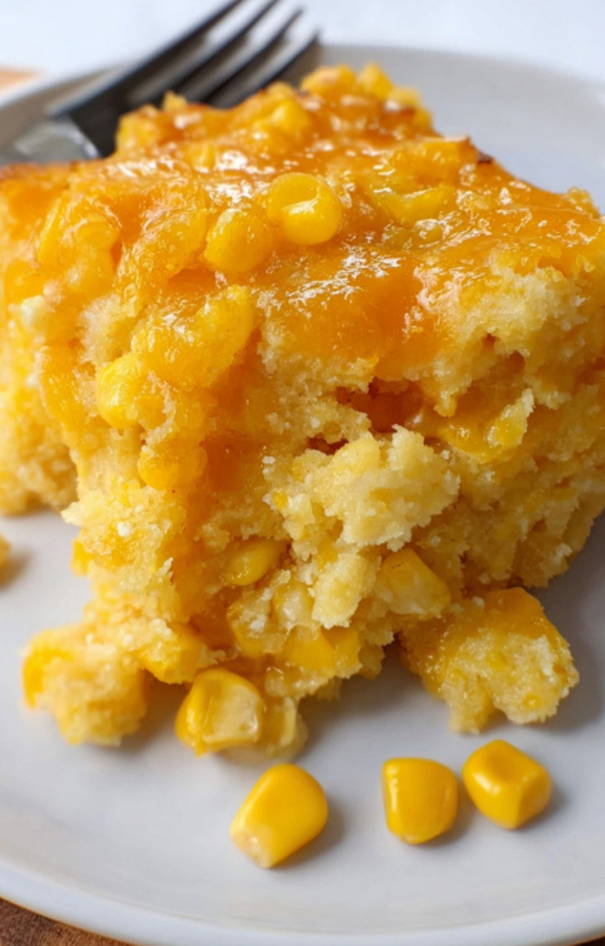 Piece of cheesy corn casserole on a plate, with sweet corn kernels and melty cheddar cheese visible
