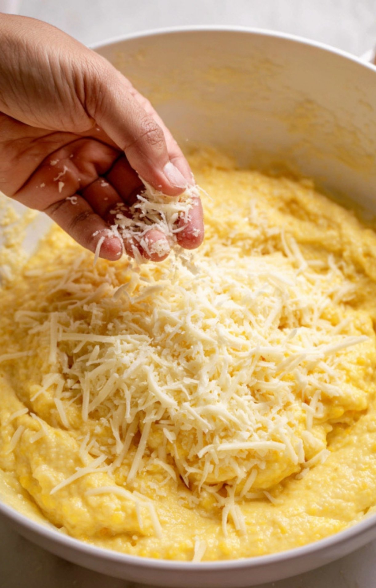 Hand adding shredded cheese to corn pudding batter in a bowl, blending creamy texture and cheesy goodness for the ultimate Southern corn casserole.