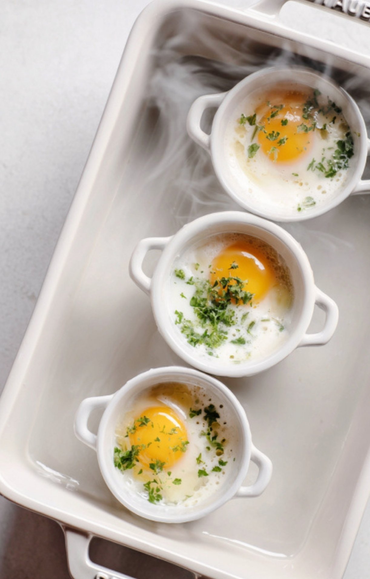 Three cocottes with eggs and herbs baking in a hot water bath, showing the gentle cooking process for soft, custardy eggs.