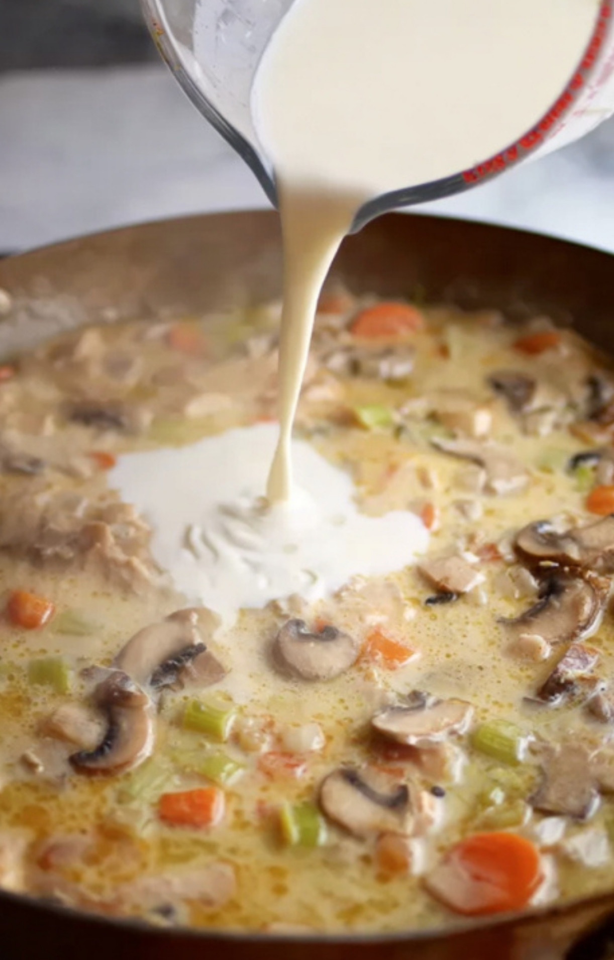 Pouring cream into a skillet filled with mushrooms, carrots, celery, and chicken.