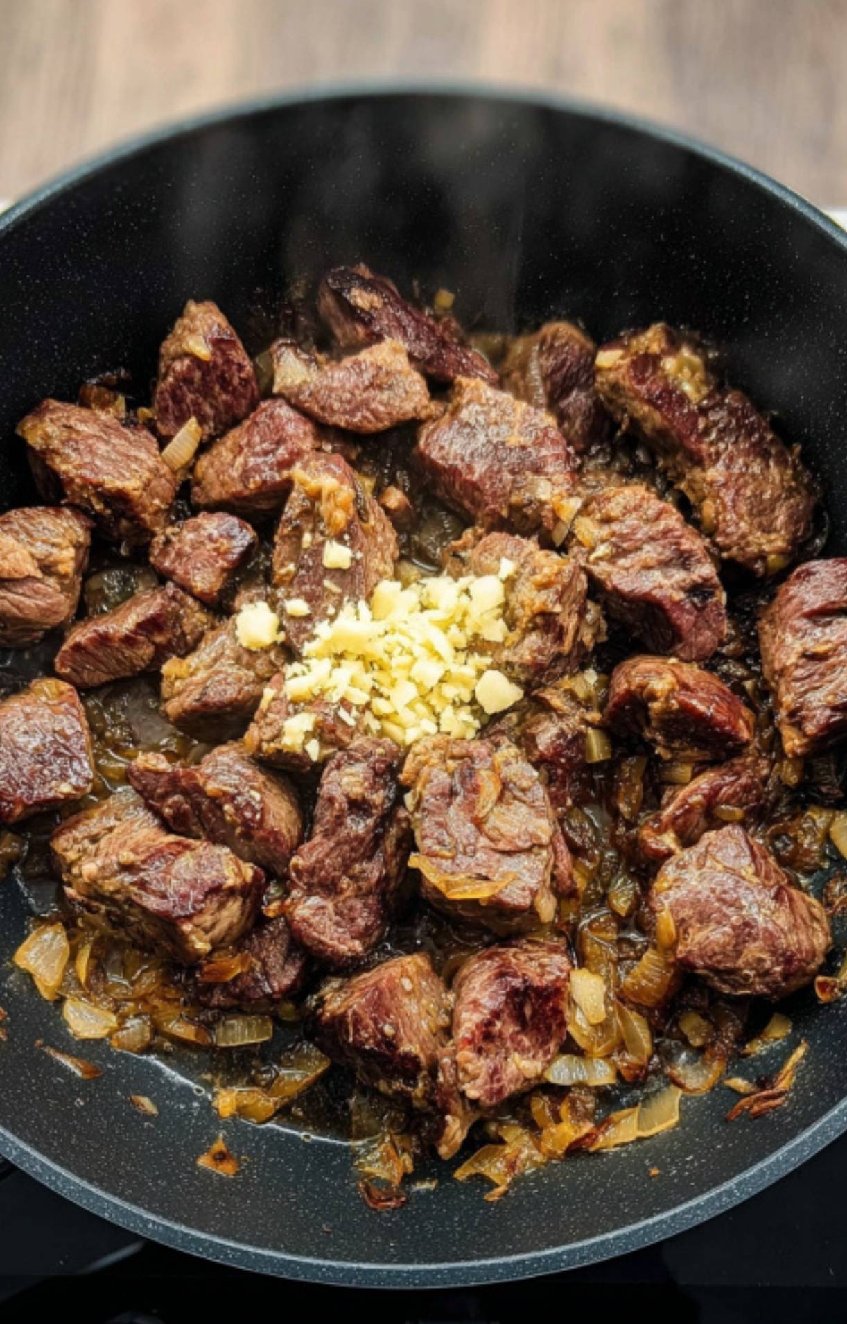 Seared beef cubes in a pan with sautéed onions and fresh minced garlic, building the savory base for slow cooker Hungarian goulash.
