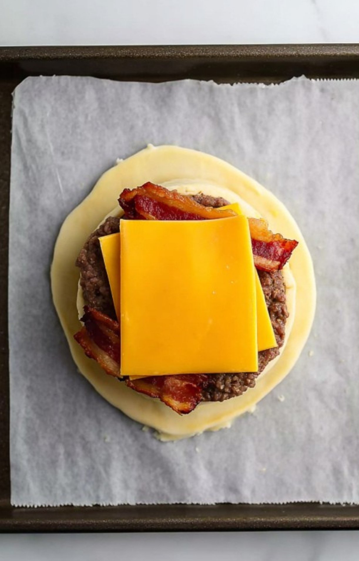 Overhead shot of ground beef, bacon, cheddar cheese, egg, butter, and puff pastry on a marble countertop. All the fresh ingredients ready for homemade Beef Wellington, perfect for a holiday or special dinner.