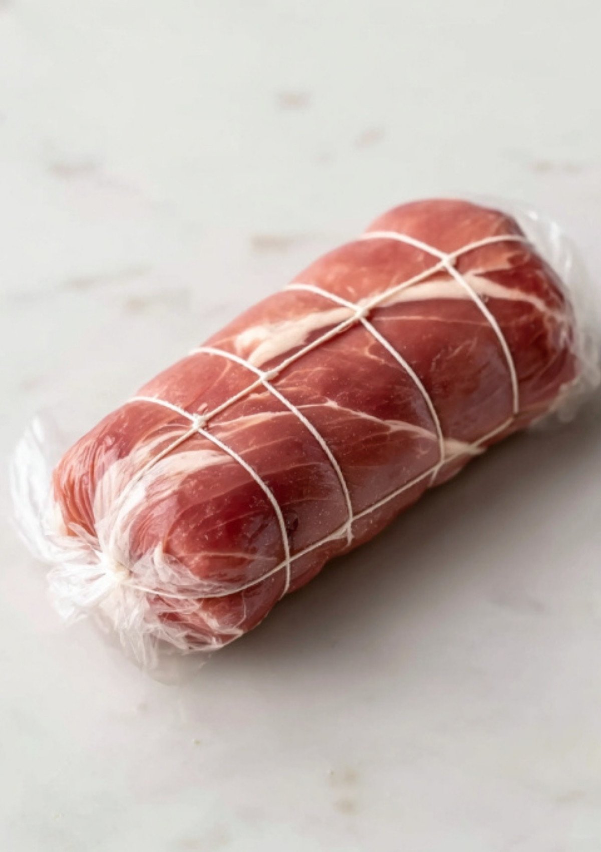 Beef fillet wrapped in prosciutto and plastic wrap, tied for shaping, as a key prep step for gluten free beef wellington recipes.