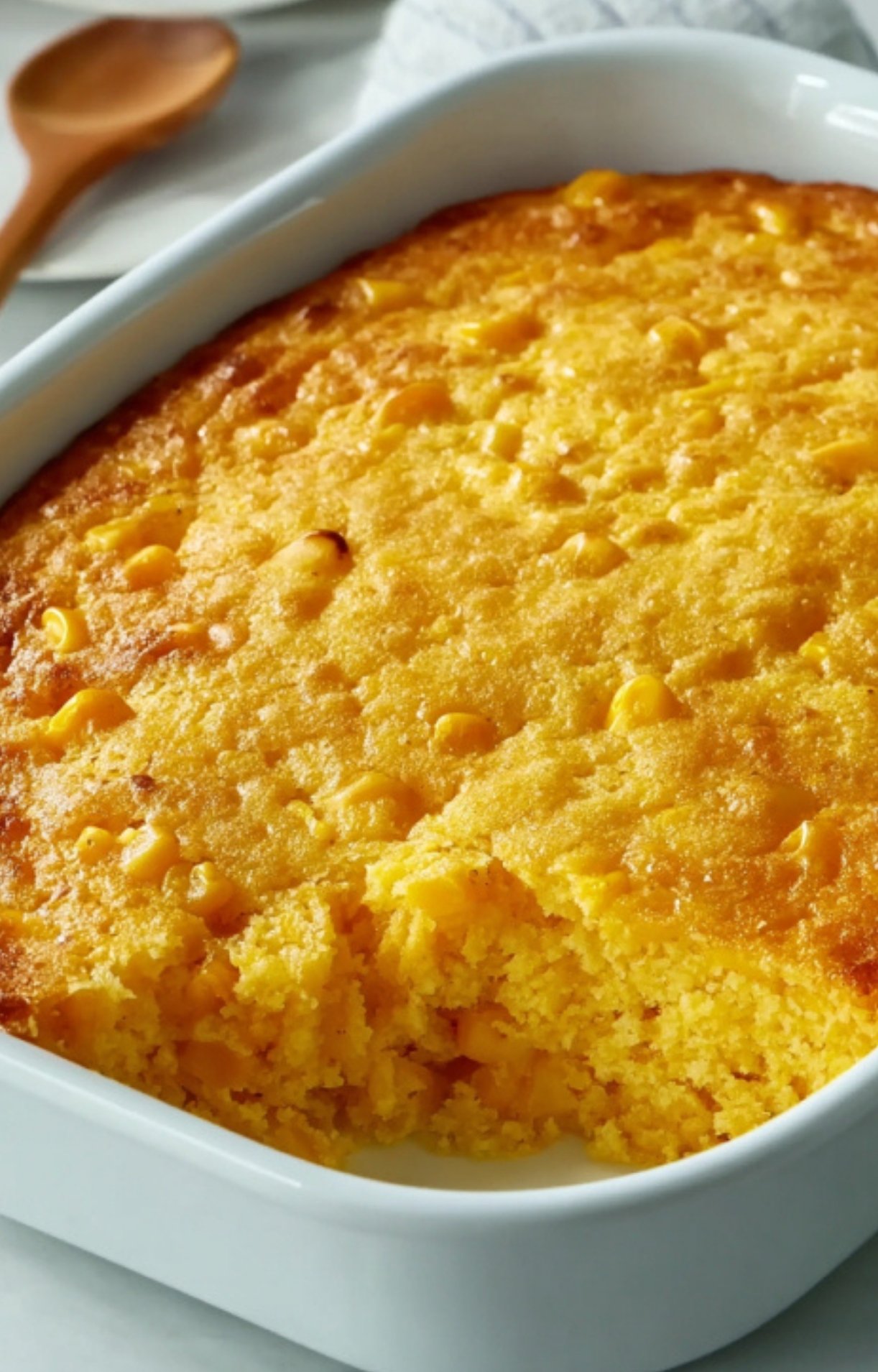 Freshly baked corn pudding casserole with a golden top and a scoop missing, showing the creamy, soft texture inside. Popular Thanksgiving and Christmas side dish.