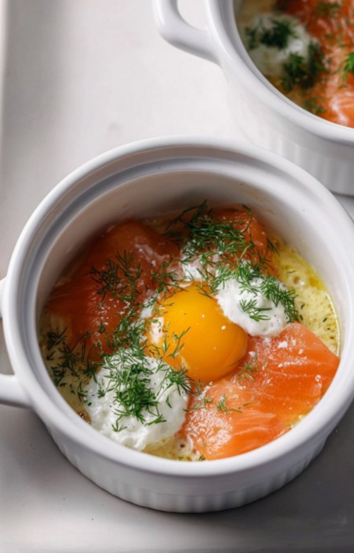 Eggs, smoked salmon, fresh dill, and dollops of cream layered in small white cocotte dishes before baking.