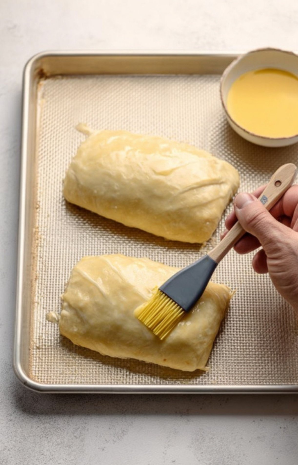 Brushing golden egg wash on pastry-wrapped ground beef Wellington before baking, for a crispy, flaky finish.