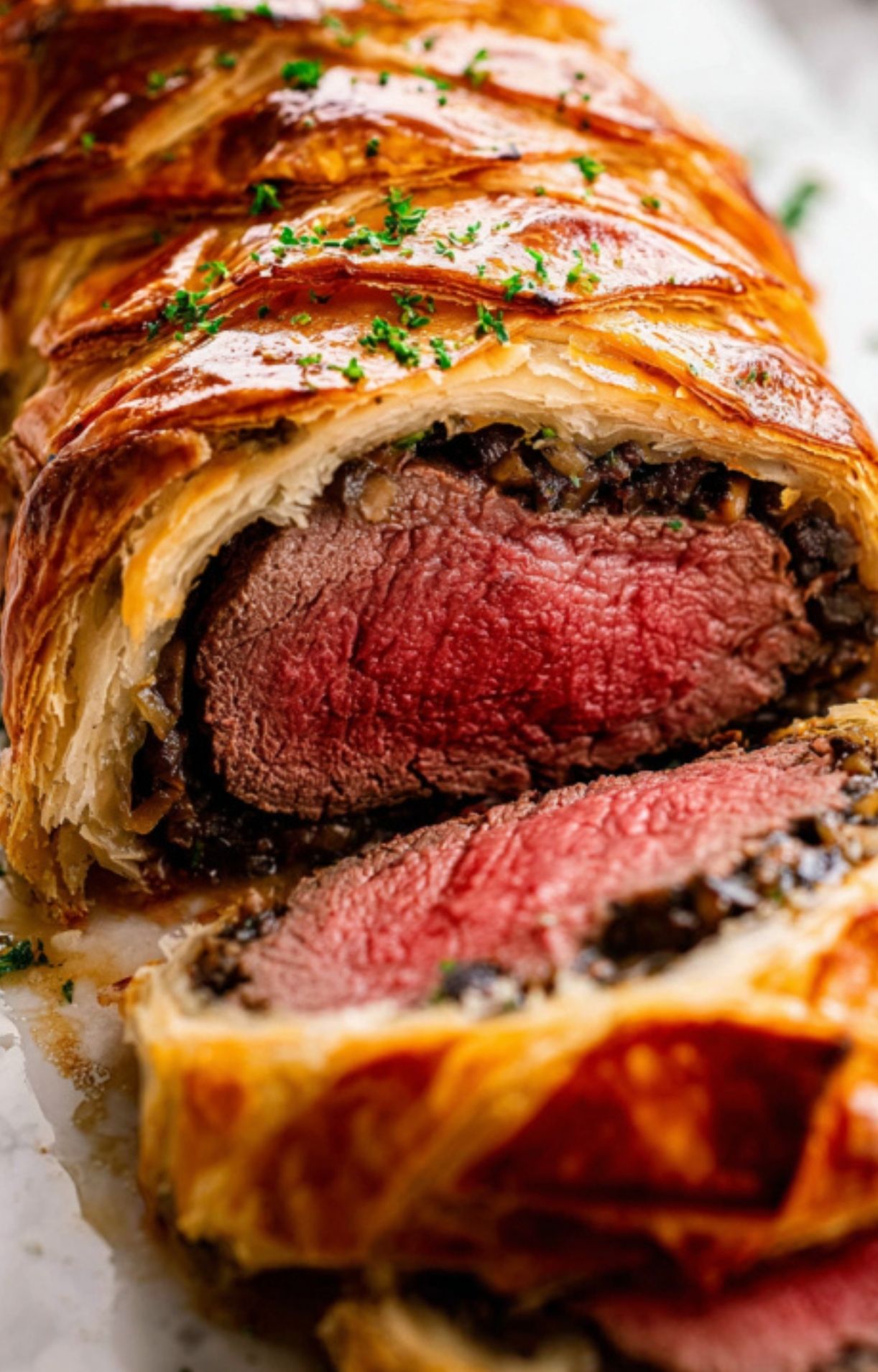 Beef wellington with several slices cut, displaying tender beef center and layers of mushroom and pastry, garnished with herbs.