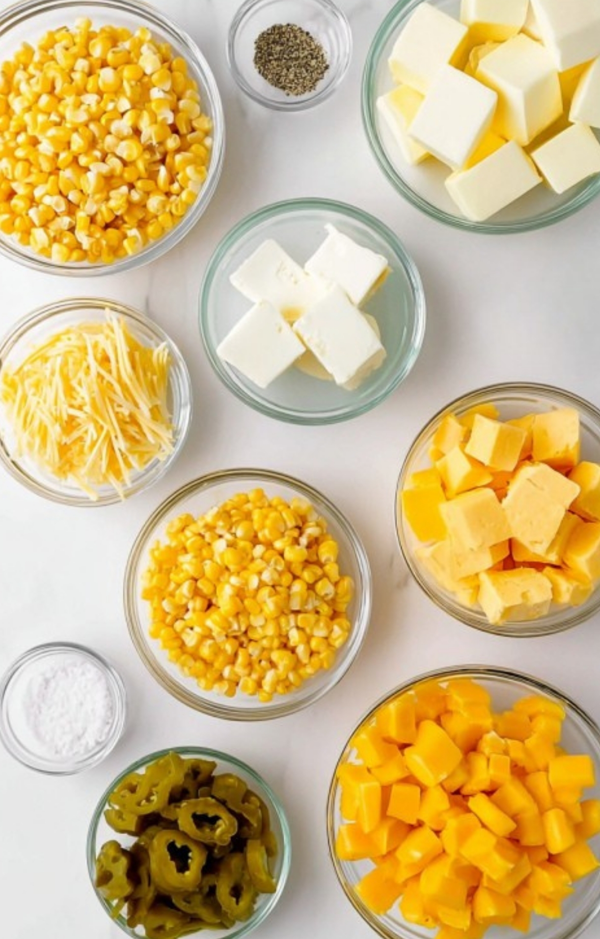 Bowls of corn, cream cheese, butter, cheese, jalapeños, and seasonings laid out on a white surface, showing all the ingredients for cream cheese corn casserole.