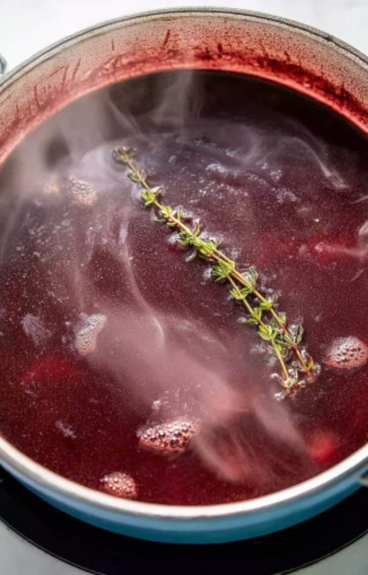 Pot of red wine reduction sauce simmering with thyme, showing the process of making a savory sauce for holiday roasts and beef wellington.
