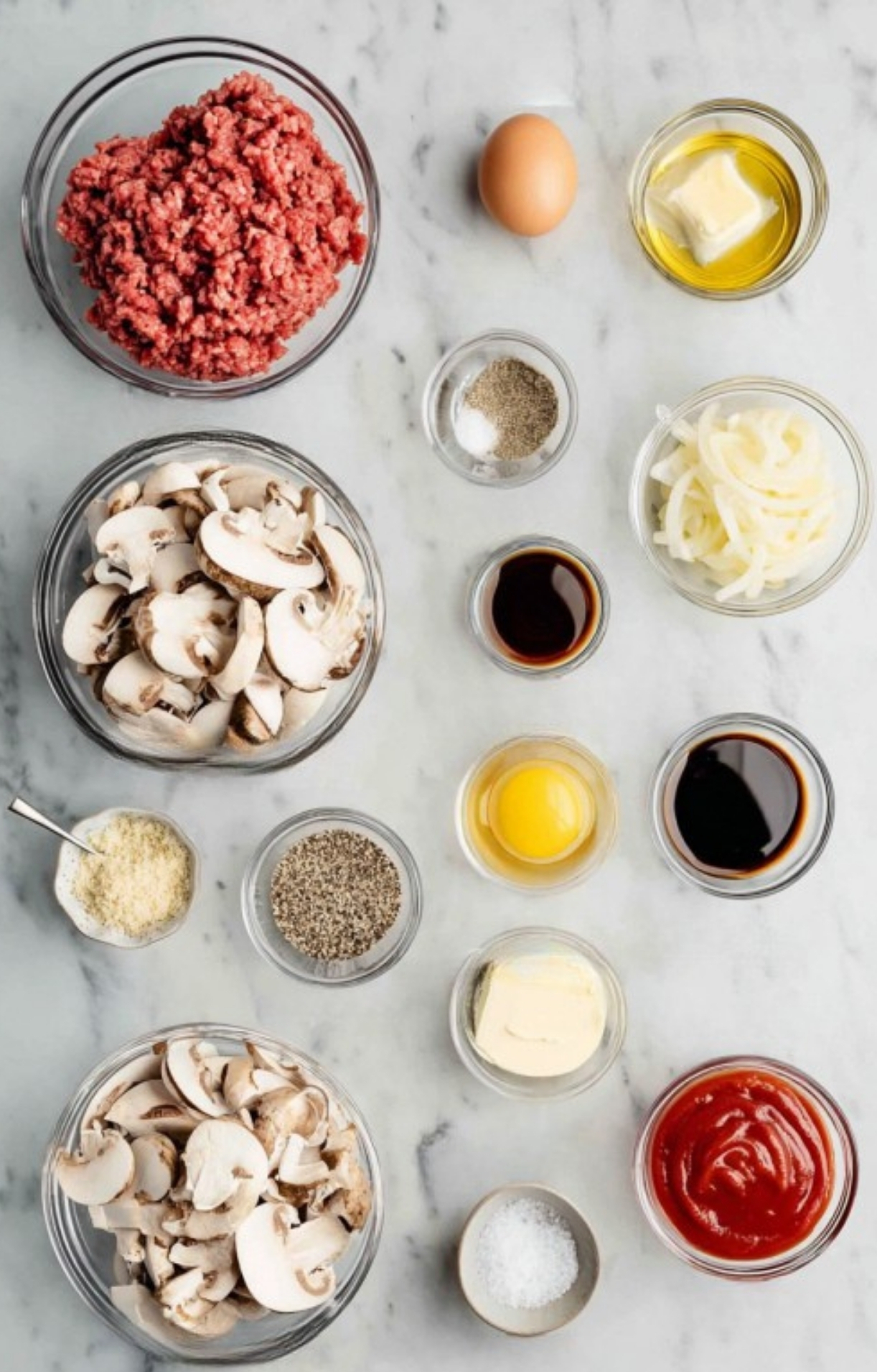 Assorted ingredients for Salisbury steak, including ground beef, eggs, onions, mushrooms, ketchup, spices, and seasonings arranged on a marble counter.