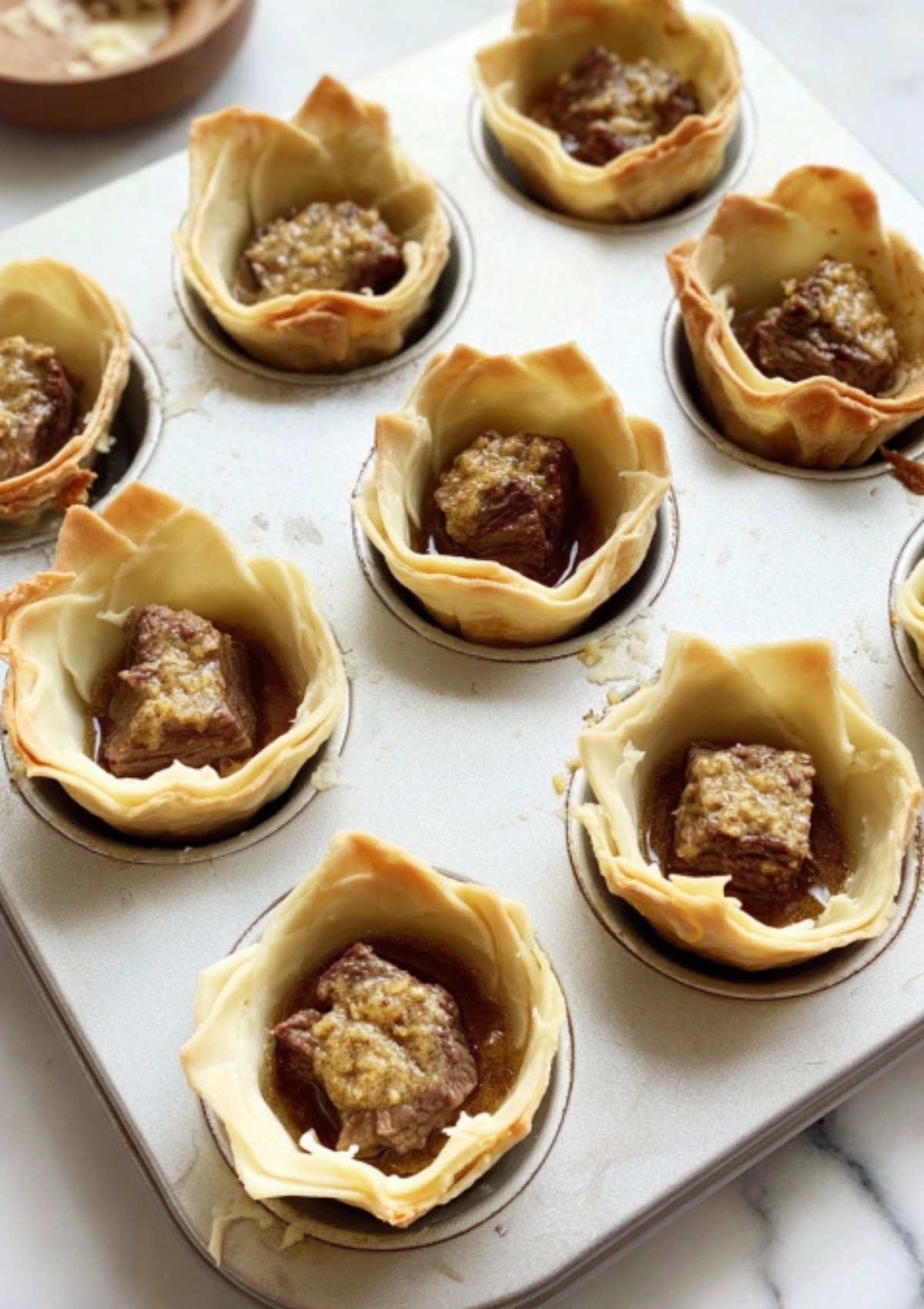 Unbaked mini beef wellingtons in muffin tin, showing beef cubes and savory filling wrapped in pastry, great for make-ahead party appetizers.